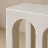 A close-up of a modern off-white concrete stool with a square top and three arched legs, set against a neutral beige background.