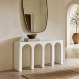 A modern white console table with five arches underneath, styled in a minimalist room with a large oval mirror, a dark wooden bowl, and books on top.