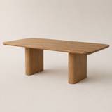 A minimalist rectangular wooden table with rounded corners and two thick block-style legs, shown on a clean, solid, warm off-white background.