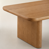 A close-up studio shot of a wooden dining table showing the corner of the tabletop with rounded edges and a thick block base, in a warm medium brown wood finish against a light beige background.