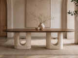 A modern oval coffee table with a dark wood top and two thick, sculptural concrete bases with U-shaped cutouts, styled in a minimalist, warm-toned interior with a vase of branches on top.