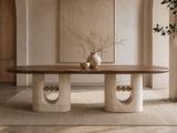 A modern oval coffee table with a dark wood top and textured stone base featuring circular cutouts and decorative gold-toned ball accents, styled in a minimalist beige interior with a vase and small bowl on top.