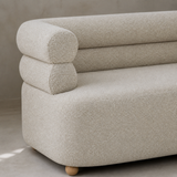 Close detail of Vernazza fine gray-ivory woven fabric showing exactly two stacked rounded bolster rails bending into one arm above the continuous seat and low base